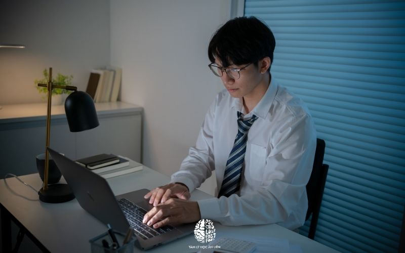 Một người trẻ ngồi bên bàn làm việc vào buổi tối, ánh sáng dịu, laptop mở nhưng người đó đang nhìn xuống sổ tay, biểu cảm trầm tĩnh, không kịch tính hóa