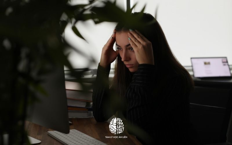 Hình ảnh một nhân viên văn phòng ôm đầu căng thẳng trước màn hình máy tính, phía sau là bóng đen của một người quản lý đang chỉ trích